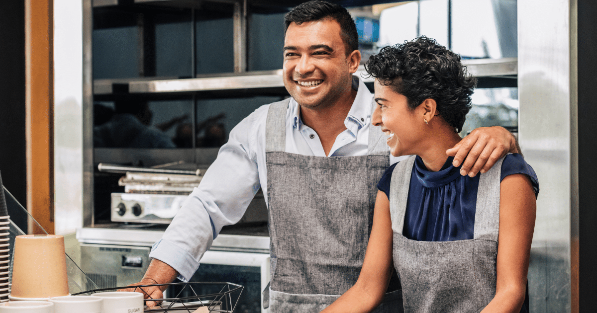 Smiling business owners in aprons discussing unsecured business loans for poor credit applicants in a cafe setting.