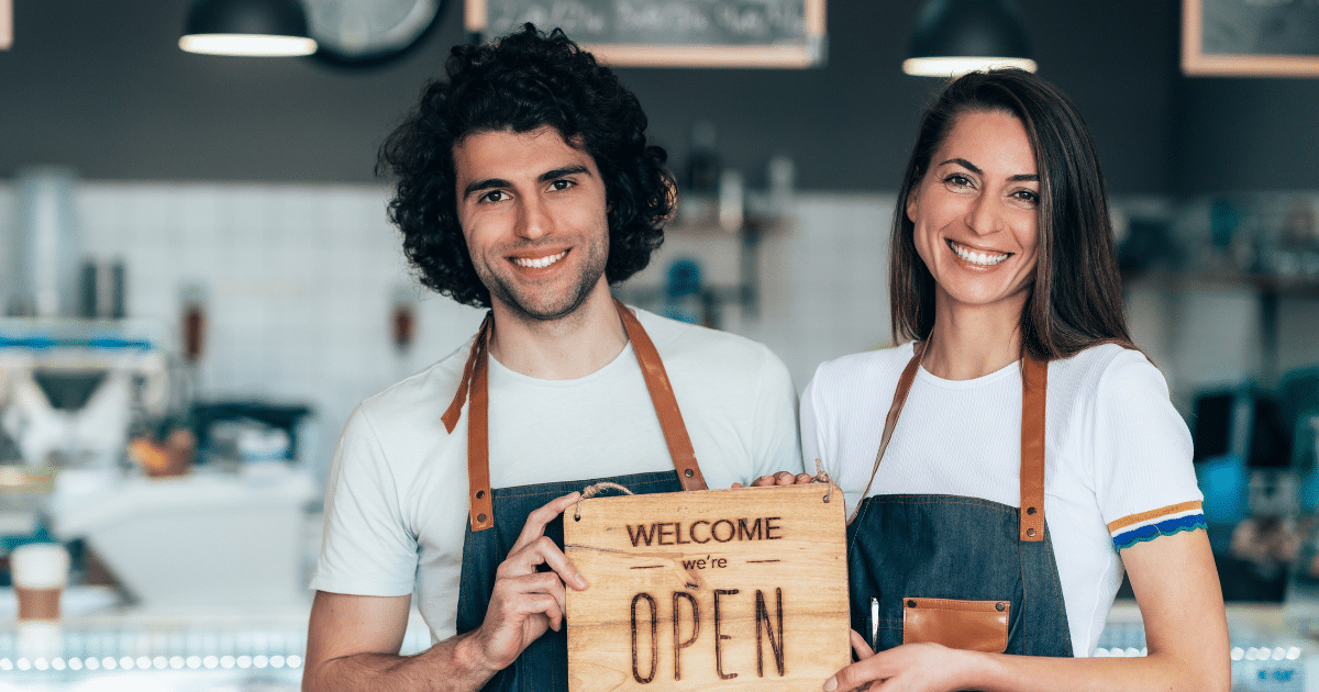 Smiling baristas holding a 'Welcome, we're OPEN' sign, showcasing excellent customer service and connection strategies.