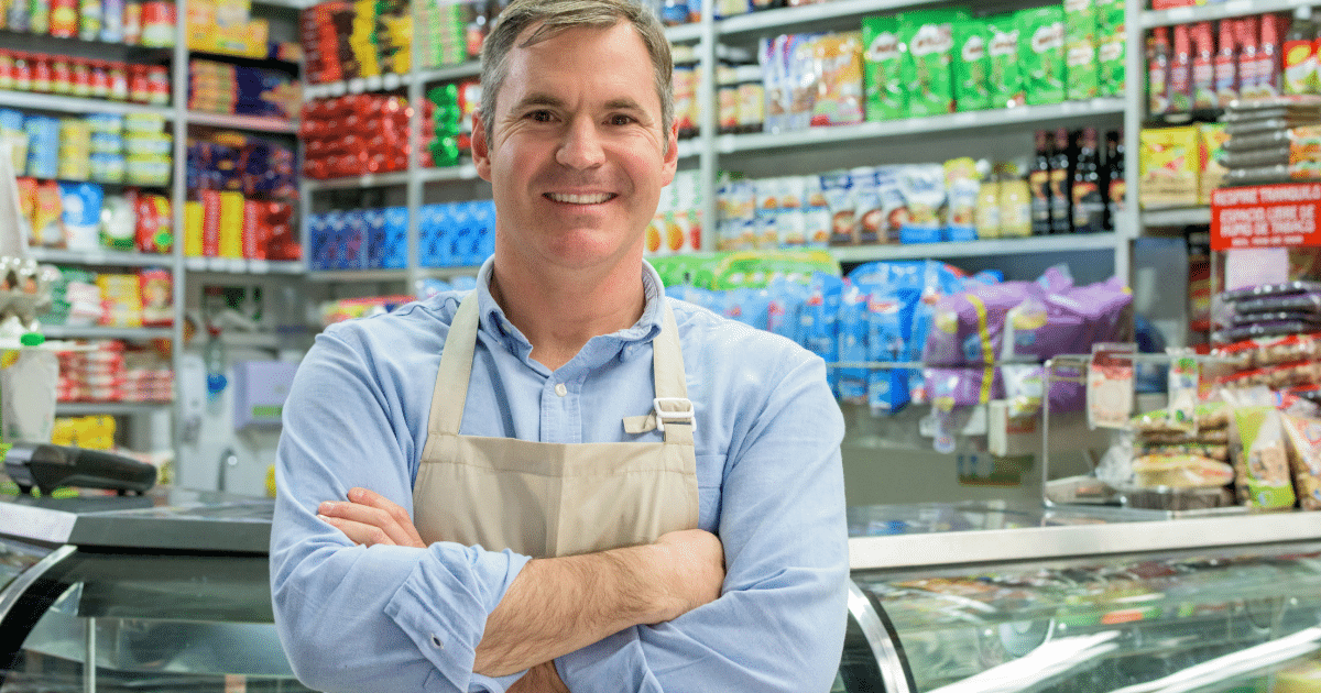 Smiling grocery store owner in apron, showcasing leadership and customer service skills in a vibrant retail environment.
