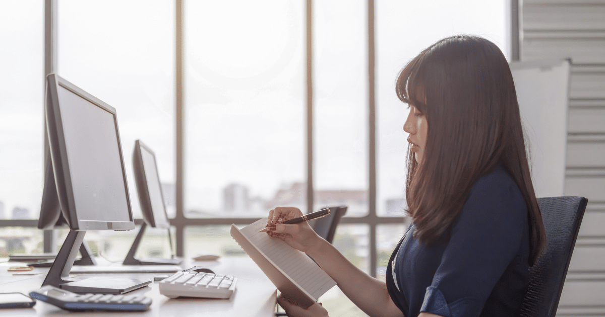 Businesswoman taking notes at a desk with computers, emphasizing profitable business habits and financial growth strategies.