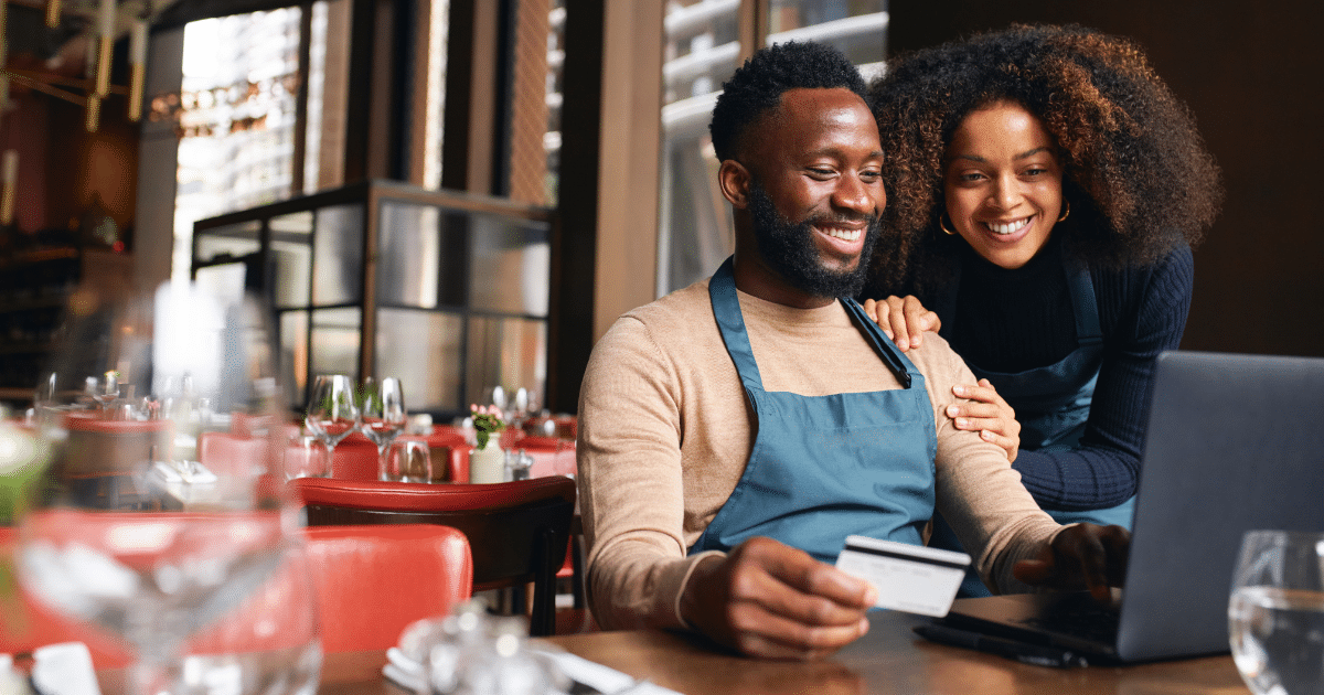 Restaurant owners exploring financing options while using a laptop and credit card in a vibrant dining setting.