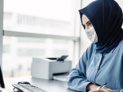 Woman in a blue shirt and mask working at a computer, representing small business growth and strategic planning.