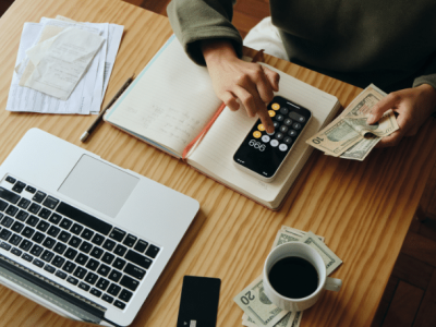 Person calculating finances with a calculator, laptop, cash, and notebook, emphasizing fiscal literacy and money management skills.
